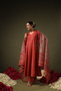 Woman in a red traditional outfit with a patterned dupatta standing against a dark background with floral decorations.