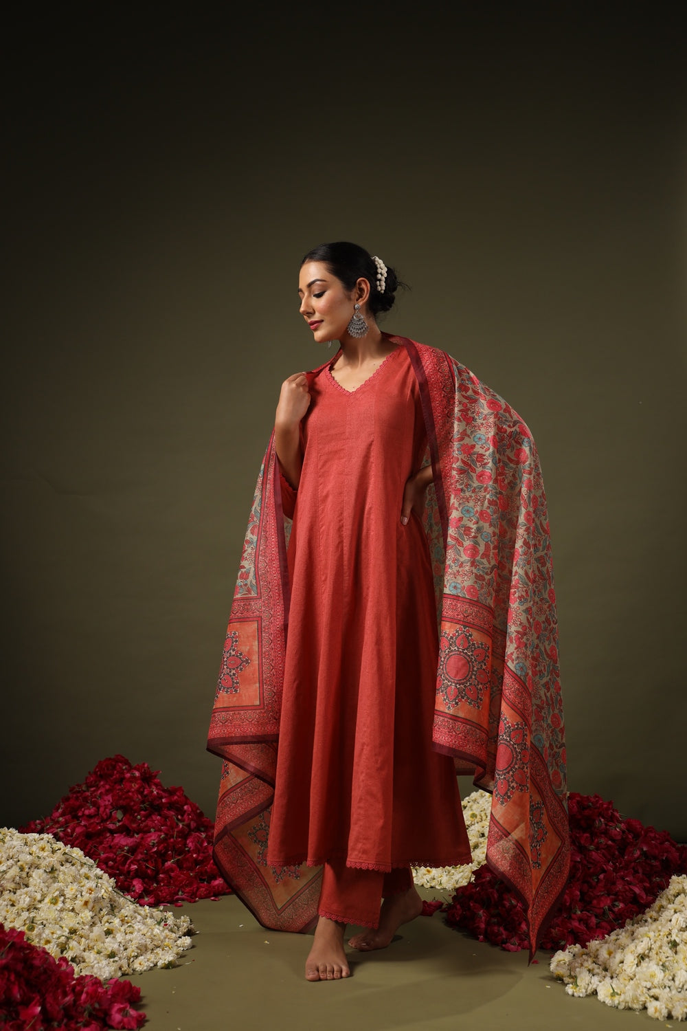 Woman in a red traditional outfit with a patterned dupatta standing against a dark background with floral decorations.