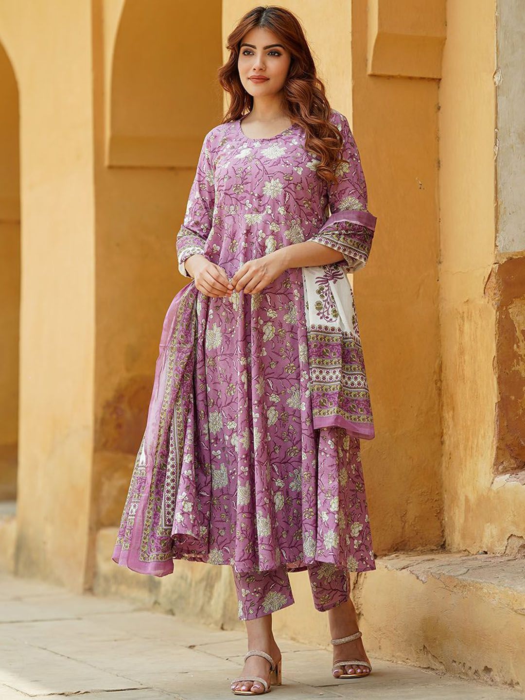 Woman wearing a purple floral outfit standing against a beige wall.