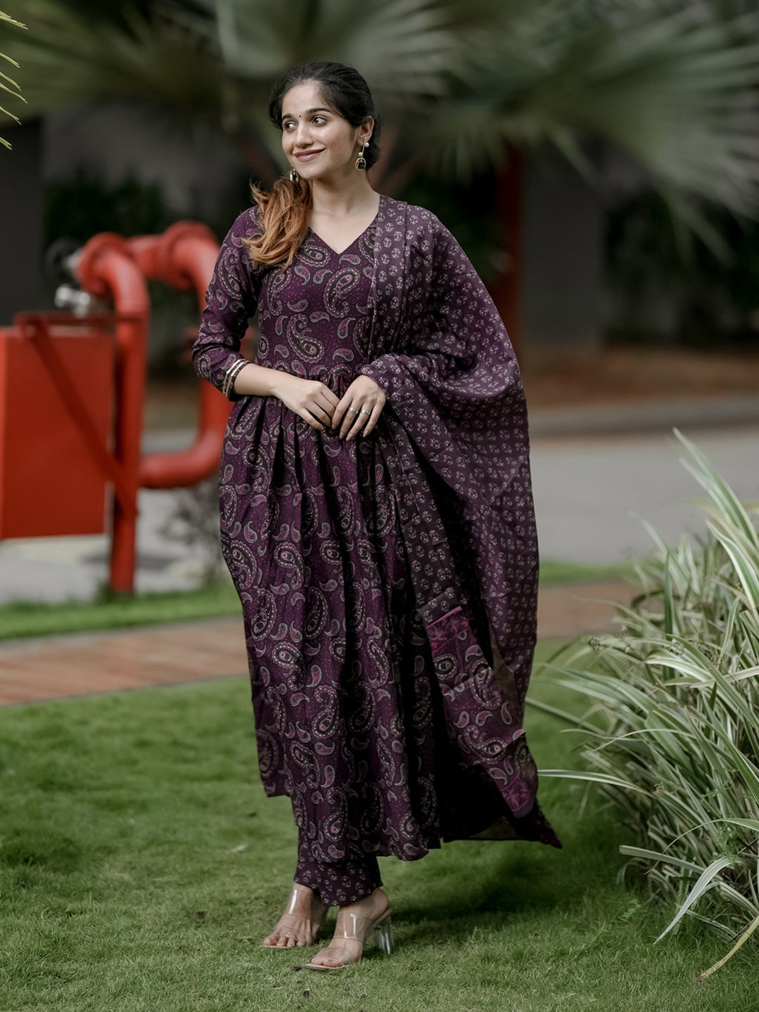 Woman in a purple patterned outfit standing outdoors with greenery in the background