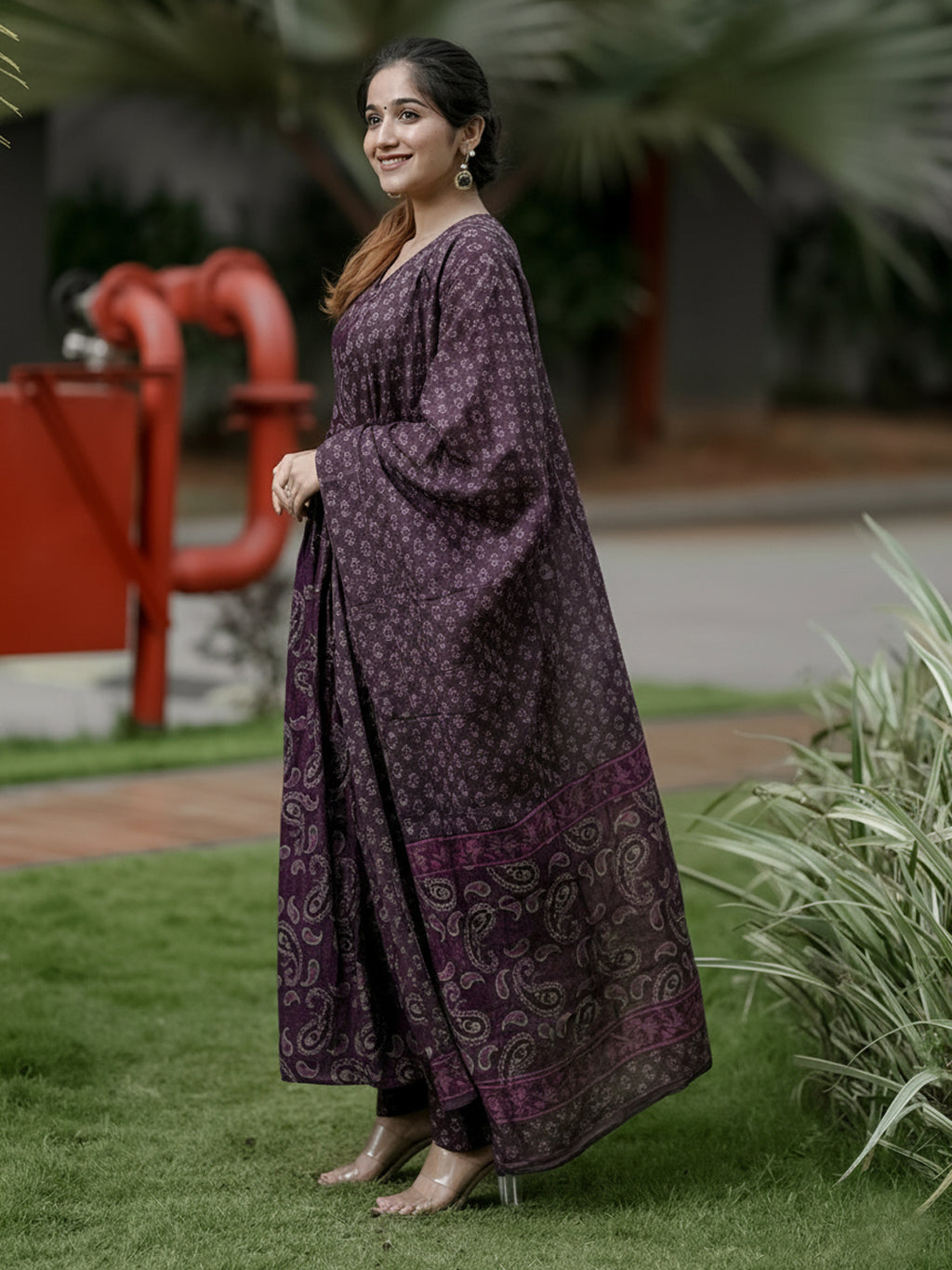 Woman in a long, dark purple patterned dress standing outdoors with greenery and a red structure in the background.