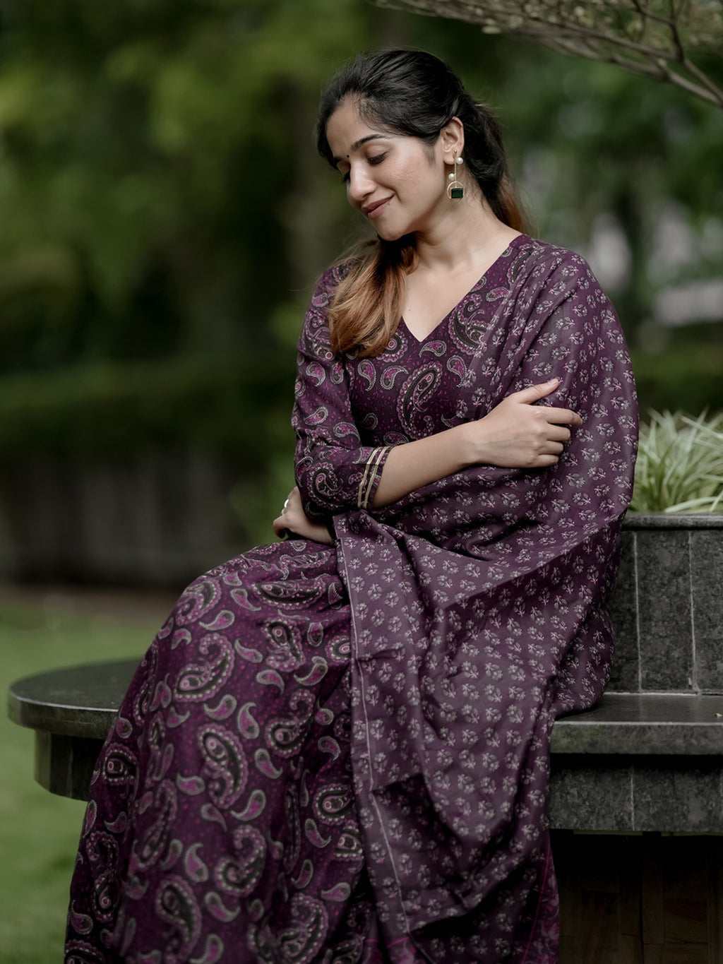 Woman in a purple patterned outfit sitting outdoors with greenery in the background