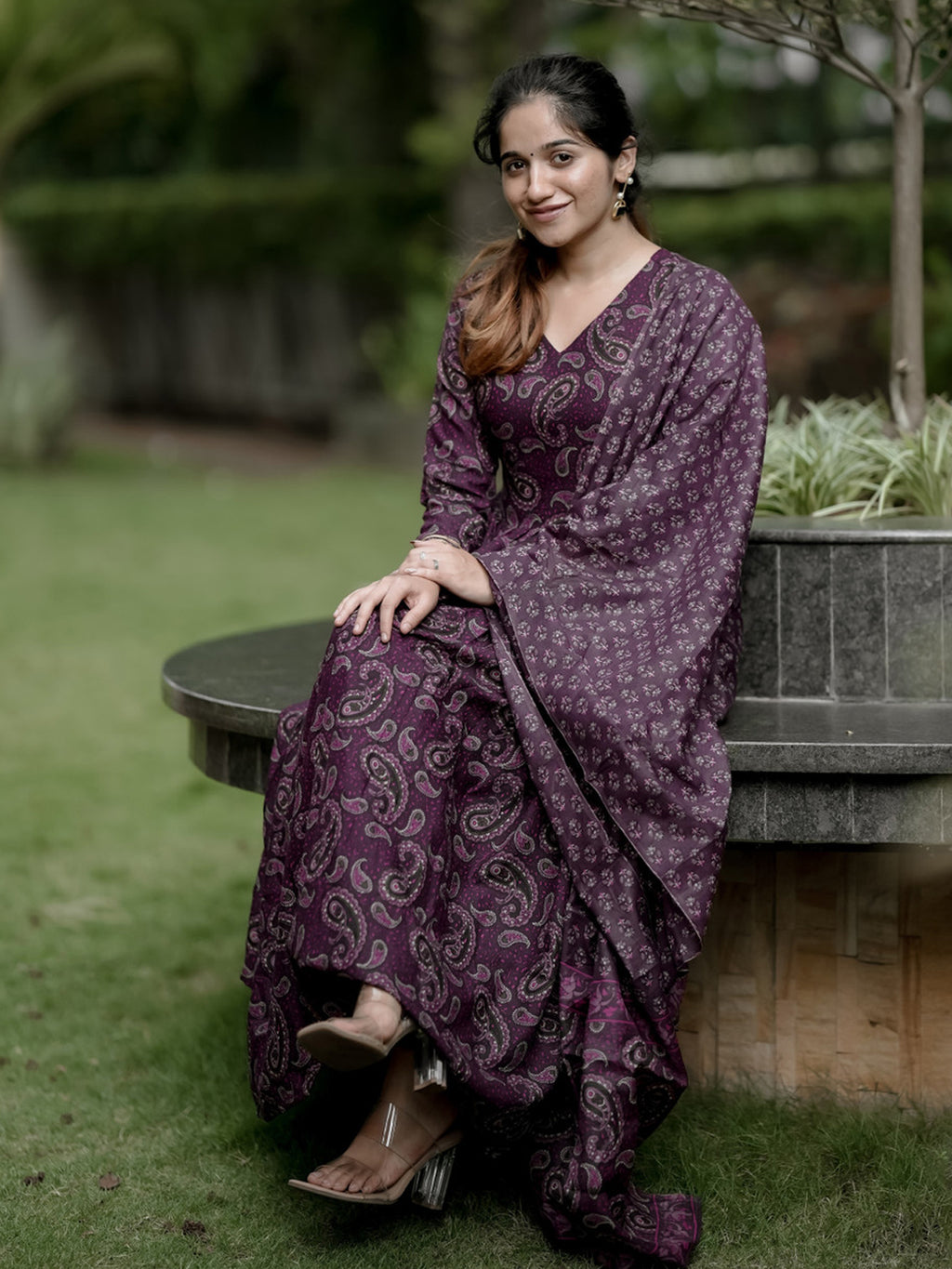 Woman in a purple patterned dress sitting on a stone bench outdoors.