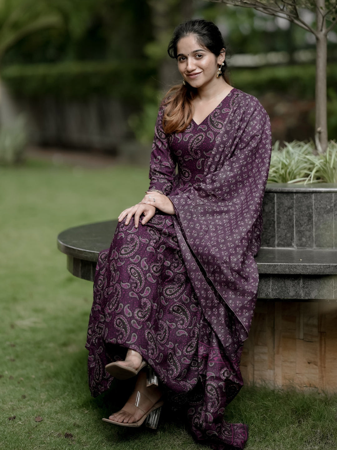 Woman in a purple patterned dress sitting on a stone bench outdoors.