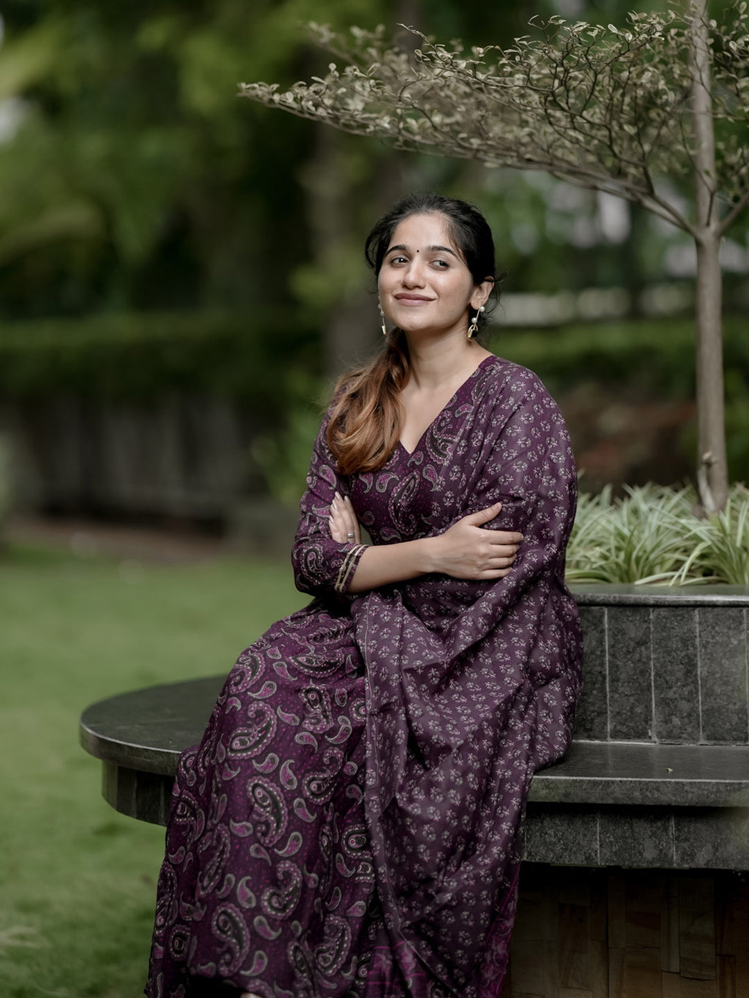 Woman in a purple patterned dress sitting outdoors with greenery in the background