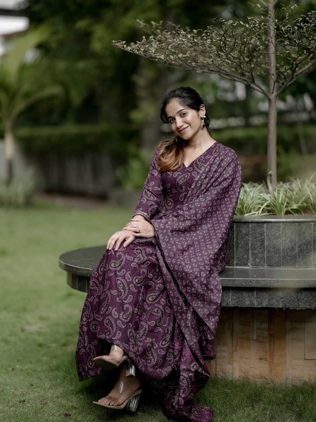 Woman in a purple saree sitting outdoors near a planter with greenery.