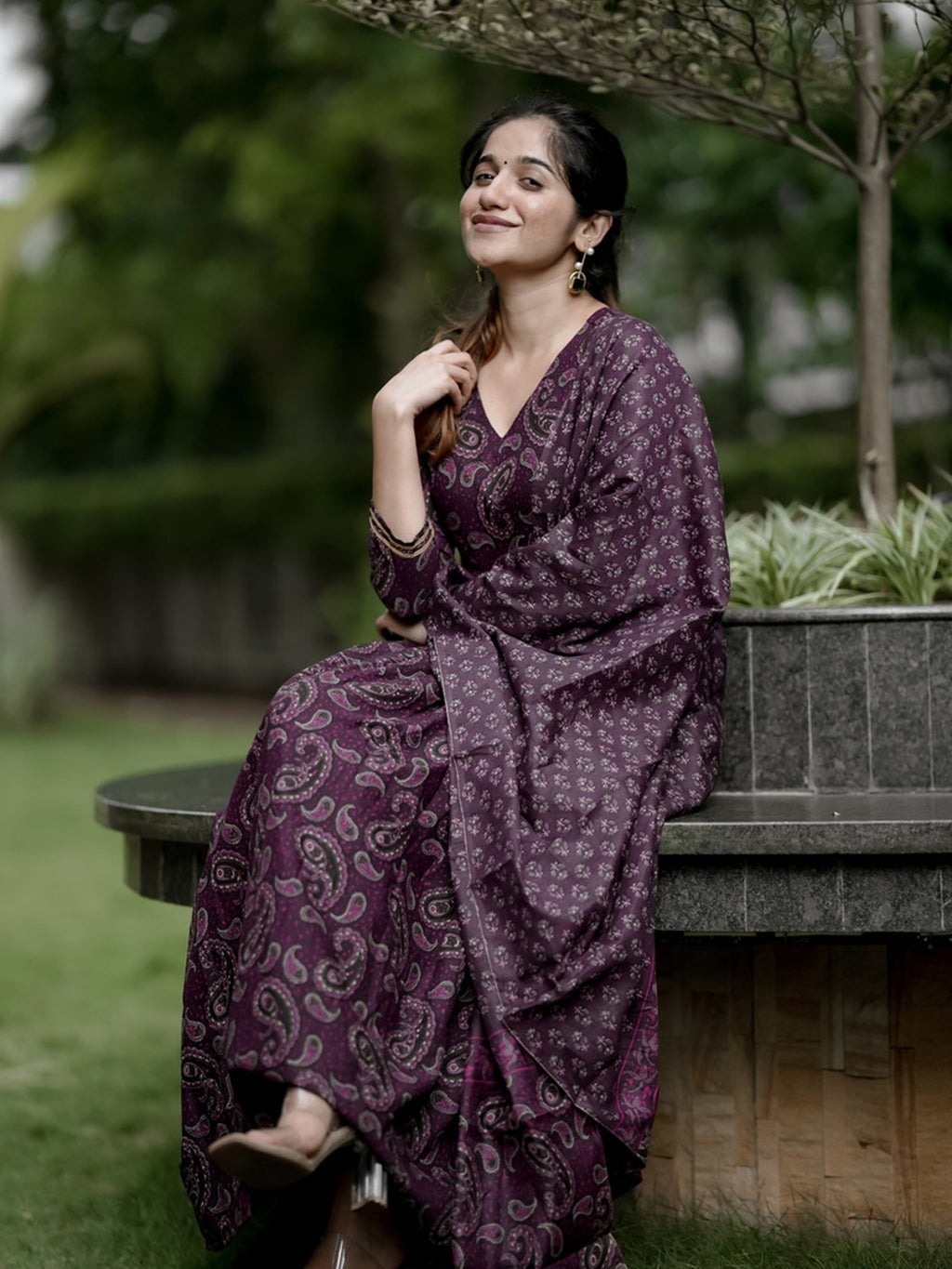 Woman in a purple patterned dress sitting outdoors on a bench.