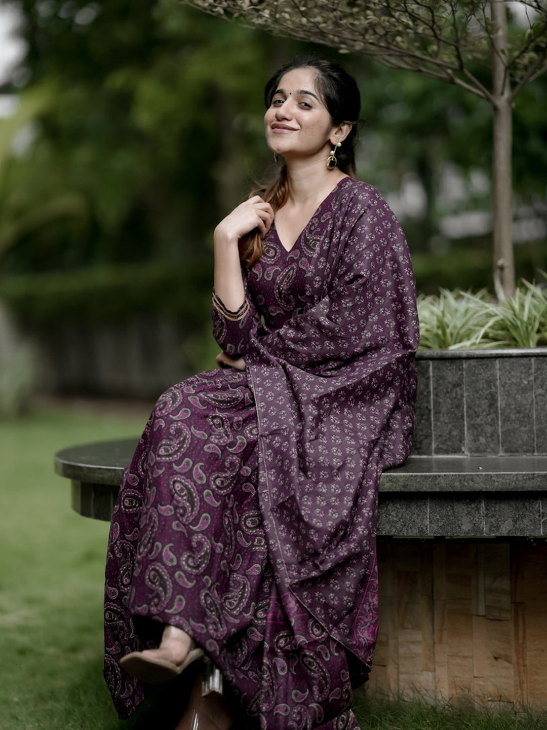 Woman in a purple patterned dress sitting outdoors on a bench.