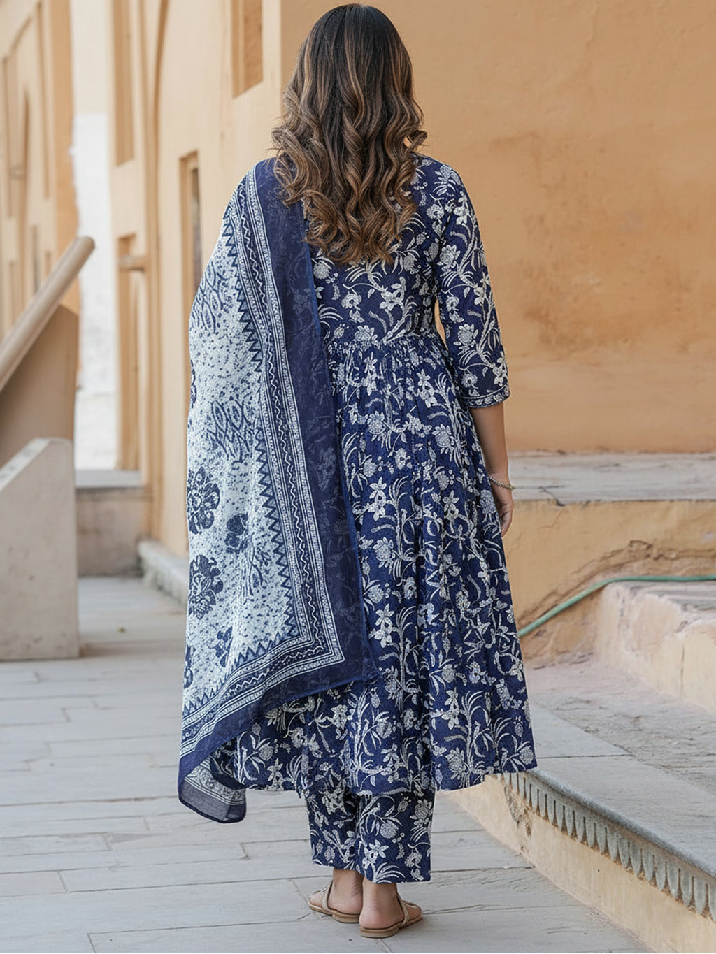 Woman in a blue floral dress and dupatta standing in front of a beige wall.