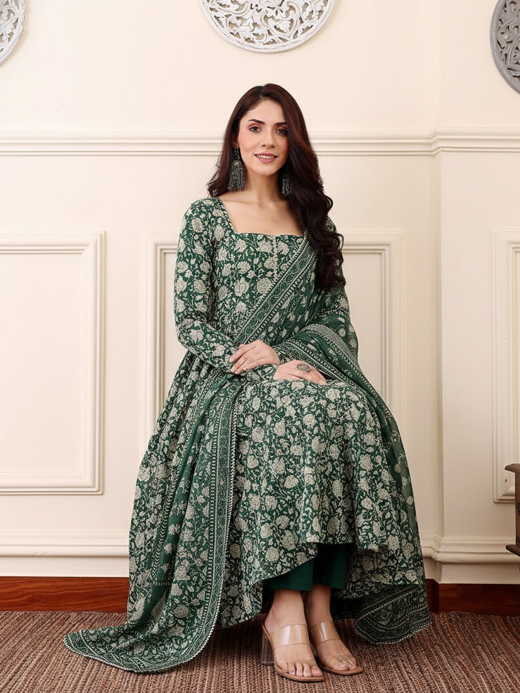 Woman in a green floral traditional outfit sitting on a wooden floor with a white wall background